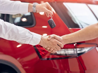 Close-up of a car salesman shaking hands with a customer and handing over car keys in front of a shiny red vehicle.